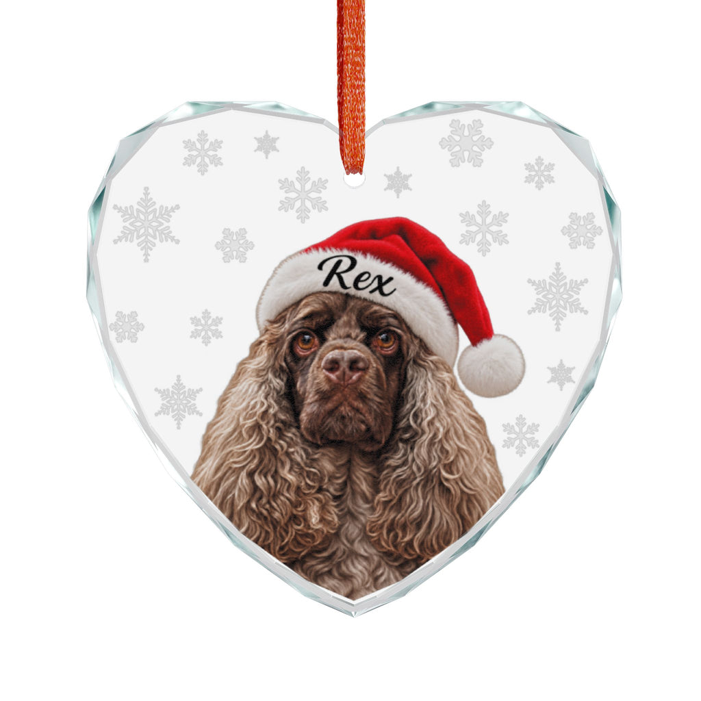A heart-shaped clear glass Christmas ornament featuring a printed portrait of a brown long-haired dog wearing a red Santa hat with white trim and the name 'Rex' on the front. The ornament has a red hanging loop at the top and a white background with faint gray snowflakes.
