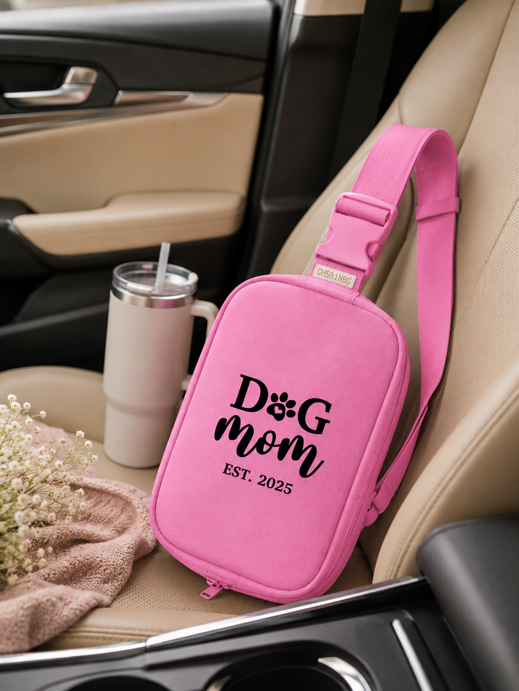 Pink bag with 'Dog Mom Est. 2025' text on a car seat next to a tumbler and flowers.