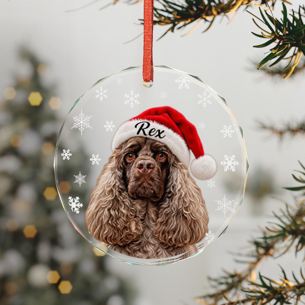 A clear round Christmas ornament hanging from a red ribbon, featuring a printed portrait of a brown dog wearing a red Santa hat with the name "Rex" on it. The ornament is decorated with white snowflakes and is photographed against a blurred Christmas tree background.