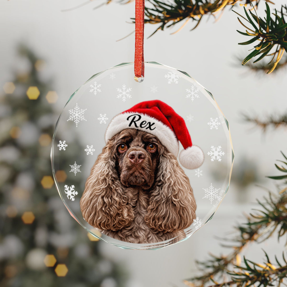 A clear round Christmas ornament hanging from a red ribbon, featuring a printed portrait of a brown dog wearing a red Santa hat with the name "Rex" on it. The ornament is decorated with white snowflakes and is photographed against a blurred Christmas tree background.