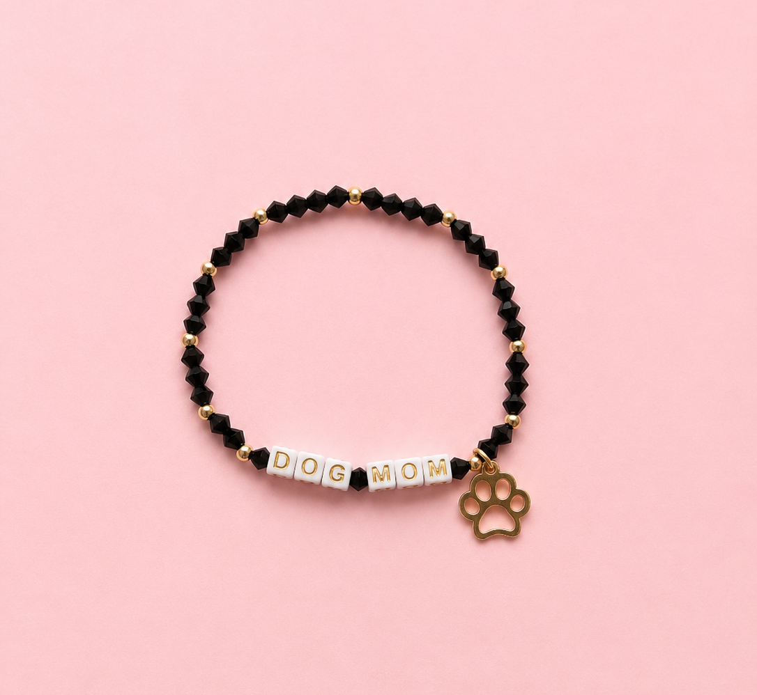 A beaded stretch bracelet featuring black faceted beads, white letter beads spelling 'DOG MOM', and a gold-tone paw print charm, displayed on a solid pink background.